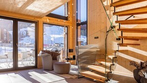 A modern interior with a wooden staircase, a glass door leading to a snowy outdoor area, and a cozy seating area with a sofa and ottoman.