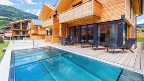 A modern wooden villa with a swimming pool, outdoor seating, and a mountain backdrop.