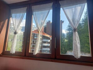 A window with three sections, each with a white lace curtain. The window overlooks a building with balconies and a mountainous landscape in the background. - Cervinia Snowy Owl (Breuil-Cervinia)