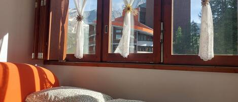 A bedroom with a bed, a window with lace curtains, and a view of buildings and mountains.