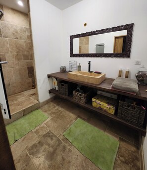 A bathroom with a stone-tiled floor, a wooden vanity with a beige sink, a large mirror, and a shower area with a tiled wall.