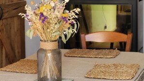 Una mesa de comedor con un jarrón de flores secas, individuales tejidos y cuadros en la pared.