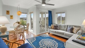 A modern living room with a grey sofa, a dining area with wicker chairs, a ceiling fan, and large windows offering a view of the outdoors. - Sands Beach Club #112 (Myrtle Beach)