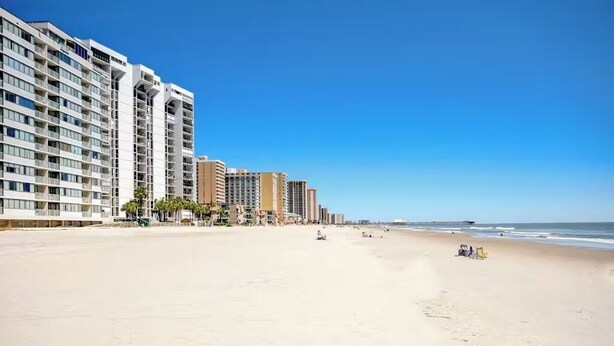 A beachfront hotel with multiple high-rise buildings, a sandy beach, and a clear blue sky. - Sands Beach Club #112 (Myrtle Beach)