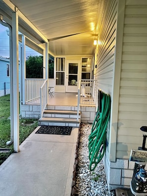 A covered porch with steps leading to a door, a small outdoor seating area, and a hose coiled against the wall.