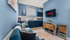 A modern living room with a sofa, a television mounted on the wall, and a kitchen area in the background.