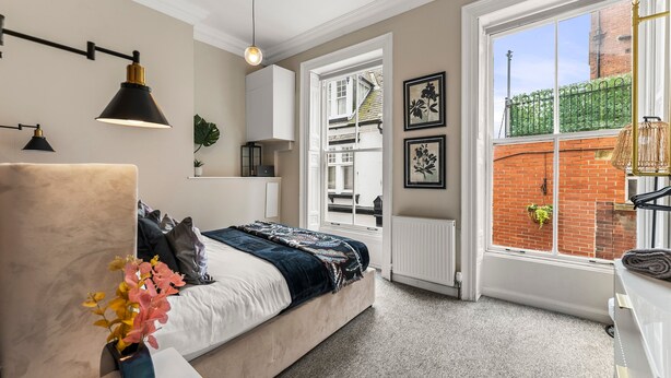 A bedroom with a bed, bedside table, lamp, window, and a view of a brick building.