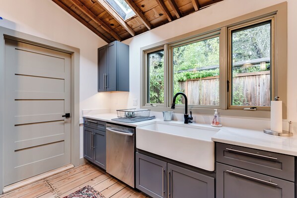 A modern kitchen with a built-in dishwasher, a large window with a view of the backyard, and a wooden ceiling.