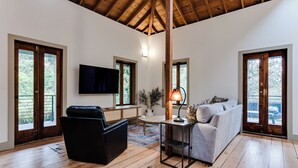 Un salón amplio con techo de madera, ventanas grandes, un sofá de cuero negro, un sofá gris y un televisor montado en la pared.