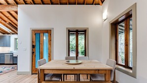 Un comedor con mesa y sillas de madera, vistas al exterior por una ventana y una cocina al fondo.