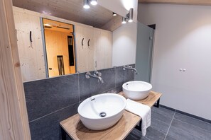 Un baño moderno con dos lavamanos blancos sobre encimeras de madera, un espejo grande y paredes con azulejos oscuros.