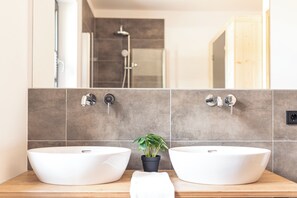 Un baño con dos lavamanos tipo sobremesa blancos, una pequeña planta en maceta y un espejo que refleja la zona de la ducha.