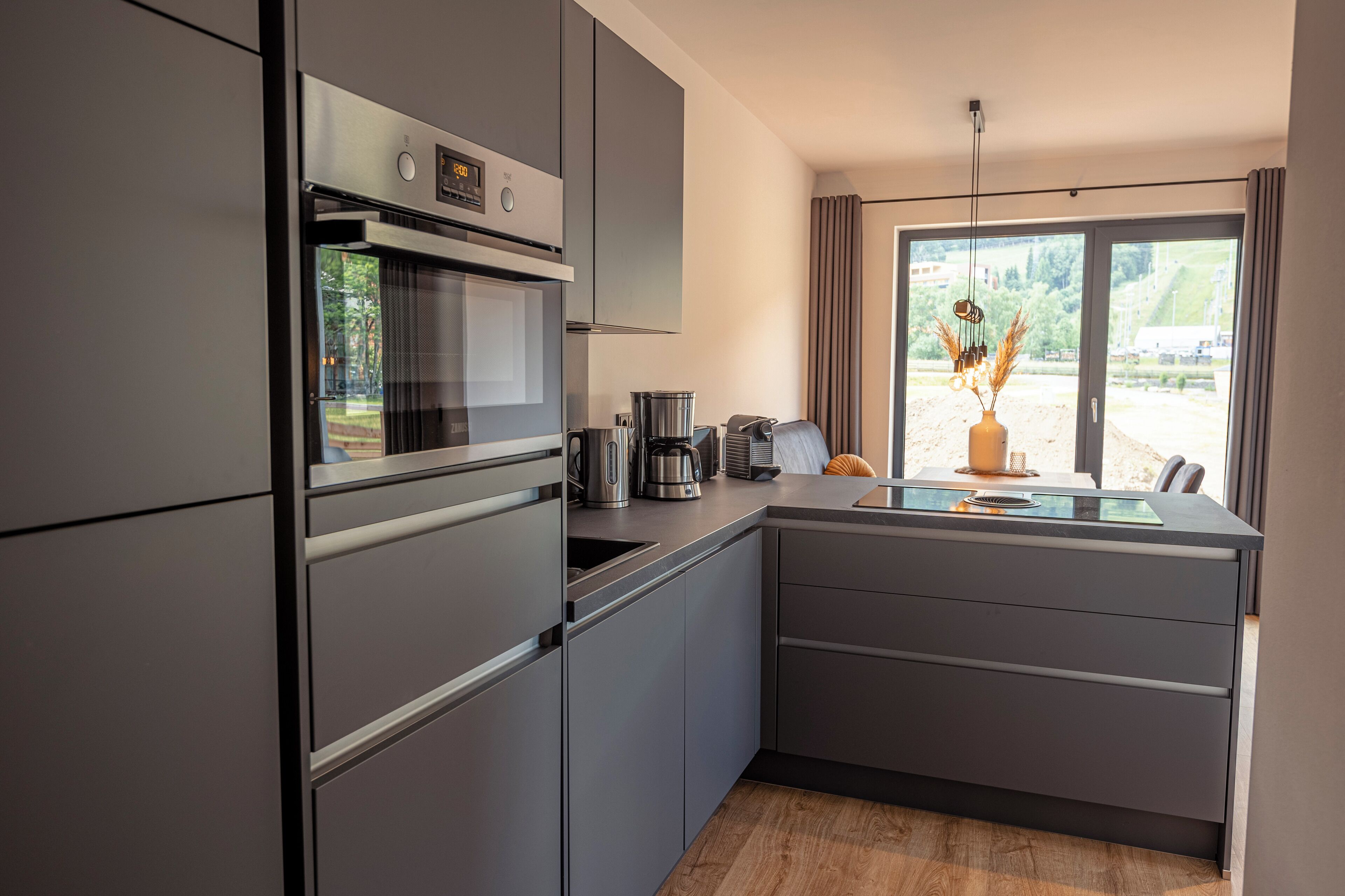 A modern kitchen with a built-in oven, a coffee maker, and a view of the outdoors through a large window.