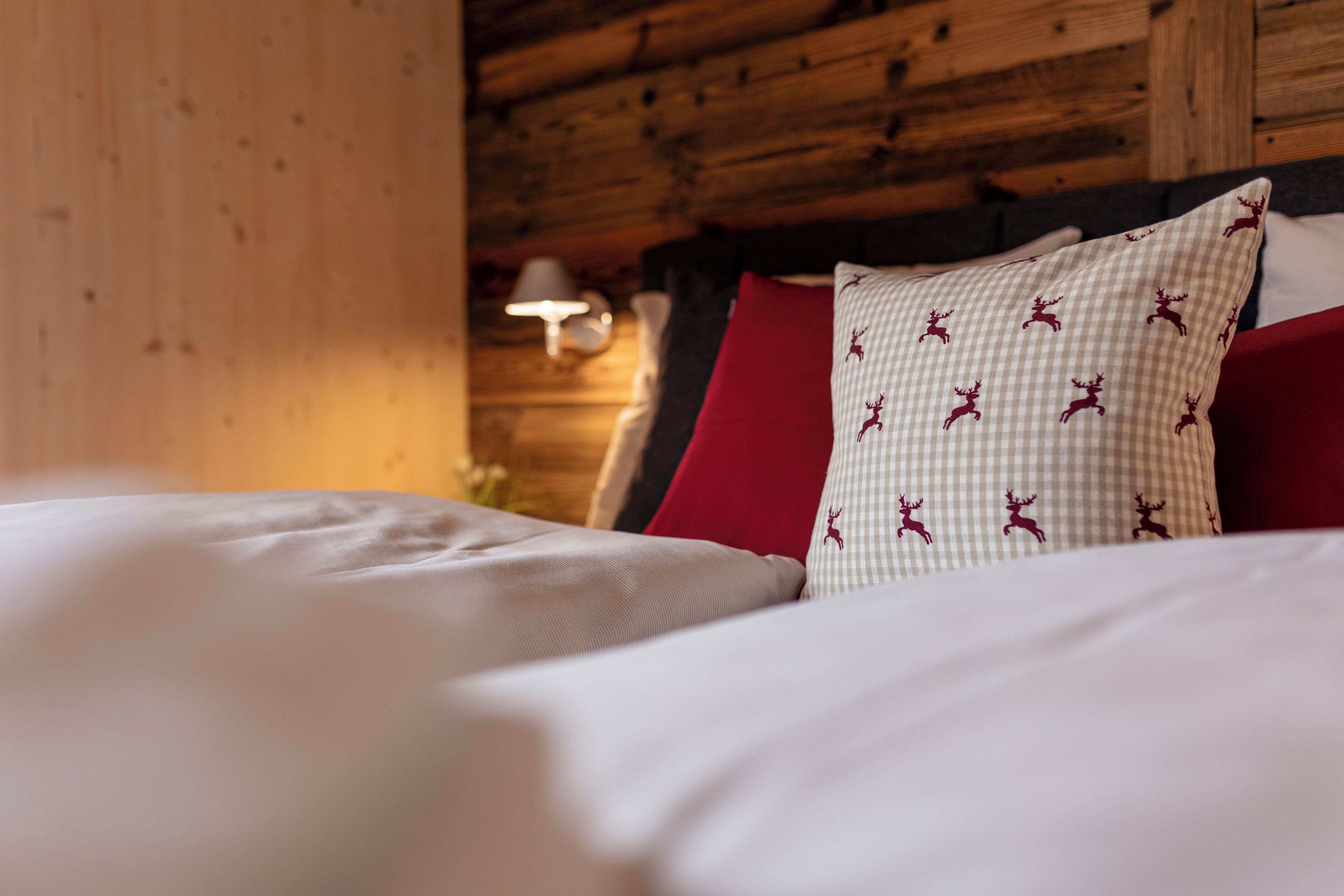 A neatly made bed with white and red bedding, a checkered pillow with reindeer patterns, and a wooden wall with a wall-mounted lamp.