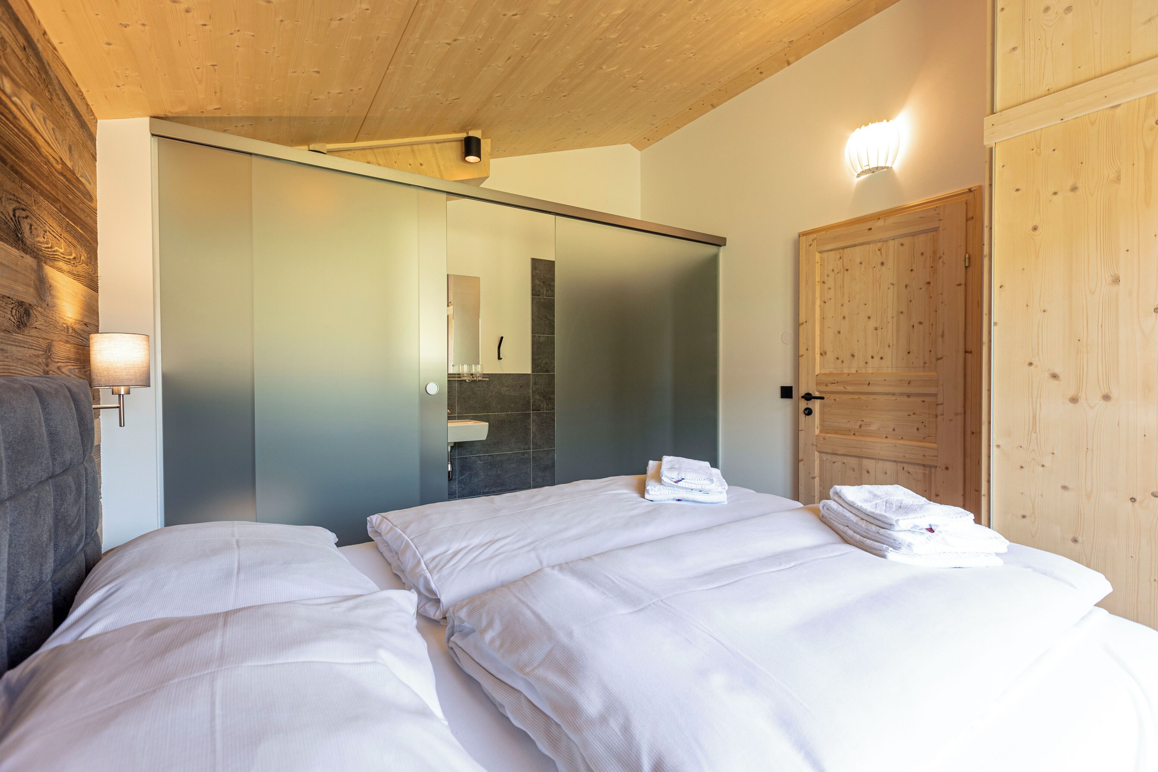 Habitación de hotel con cama, techo de madera, puerta corrediza y lámpara fijada a la pared.