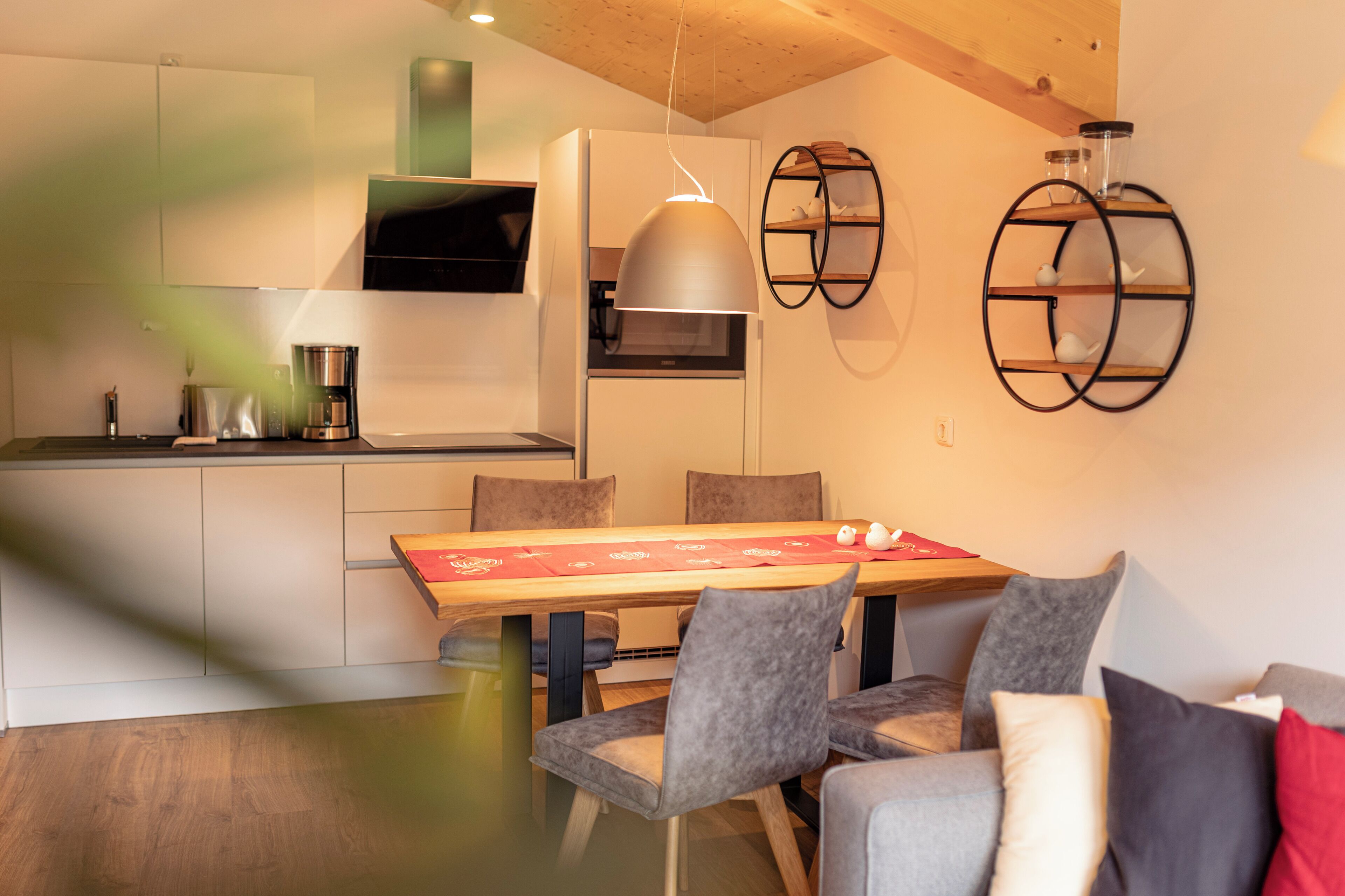 A modern kitchen with a dining area, including a wooden table with chairs, a coffee machine, and a hanging light fixture.
