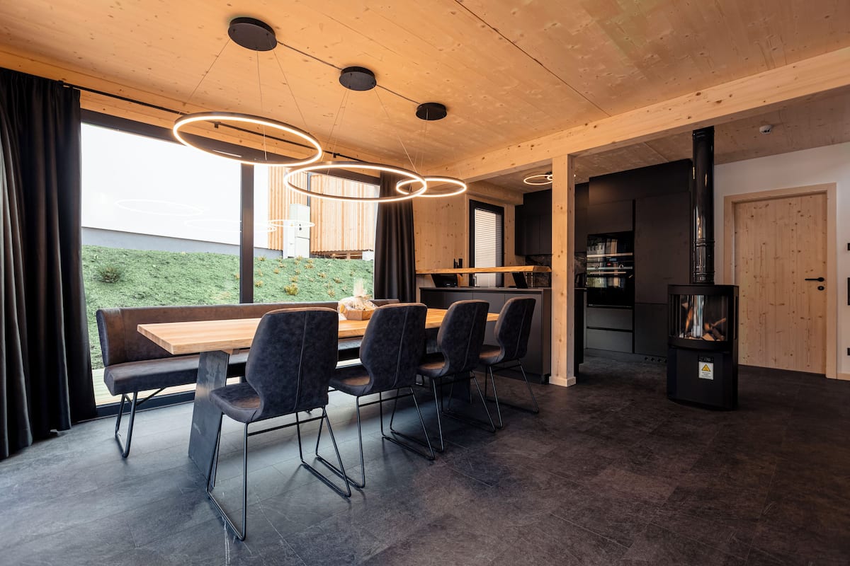 A modern dining area with a wooden table and chairs, a kitchen in the background, and a large window with curtains.