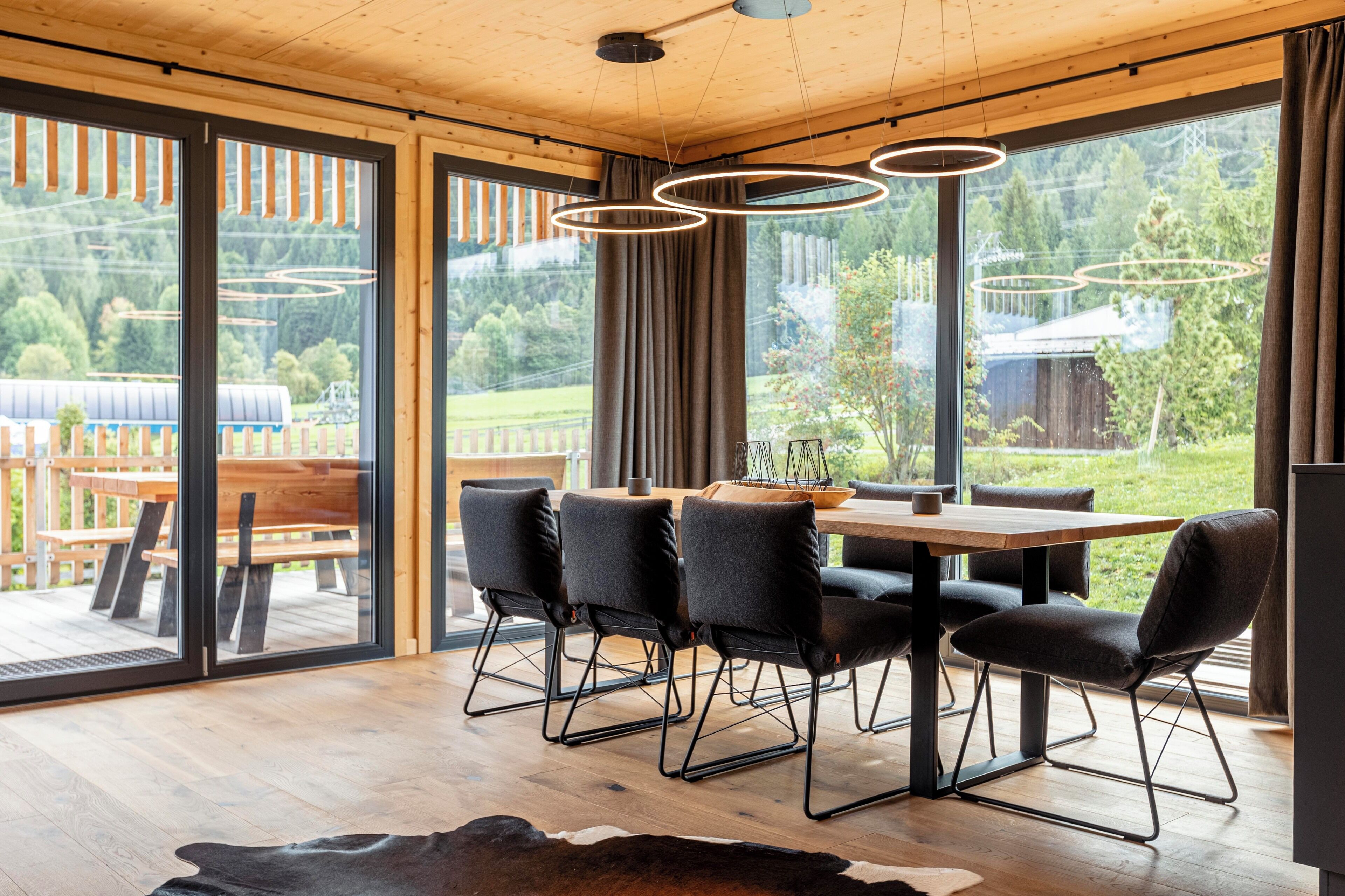 A modern dining area with a wooden table and chairs, large windows with curtains, and a view of the outdoors.