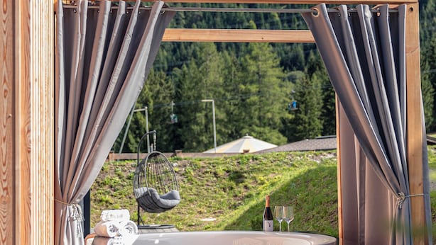 A wooden hot tub with curtains, a chair, and a bottle of wine, set against a mountainous backdrop. - Alpenchalets Biberwier by ALPS RESORTS (Biberwier)