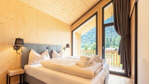 A bedroom with a large bed, wooden walls, a nightstand, and a window with a view of a wooden deck and mountains. - Alpenchalets Biberwier by ALPS RESORTS (Biberwier)