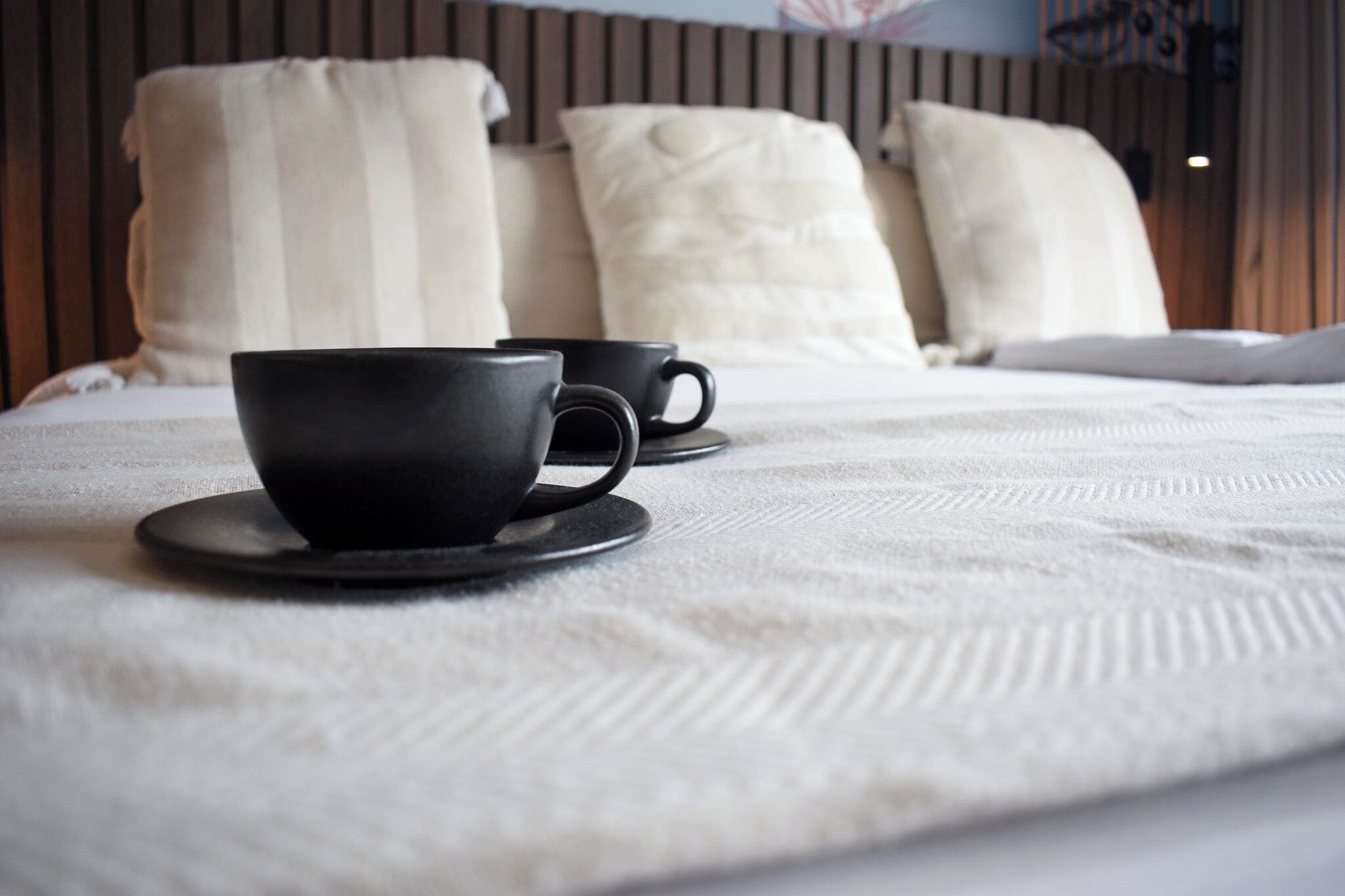 A neatly made bed with a white textured bedspread and two black cups on saucers.
