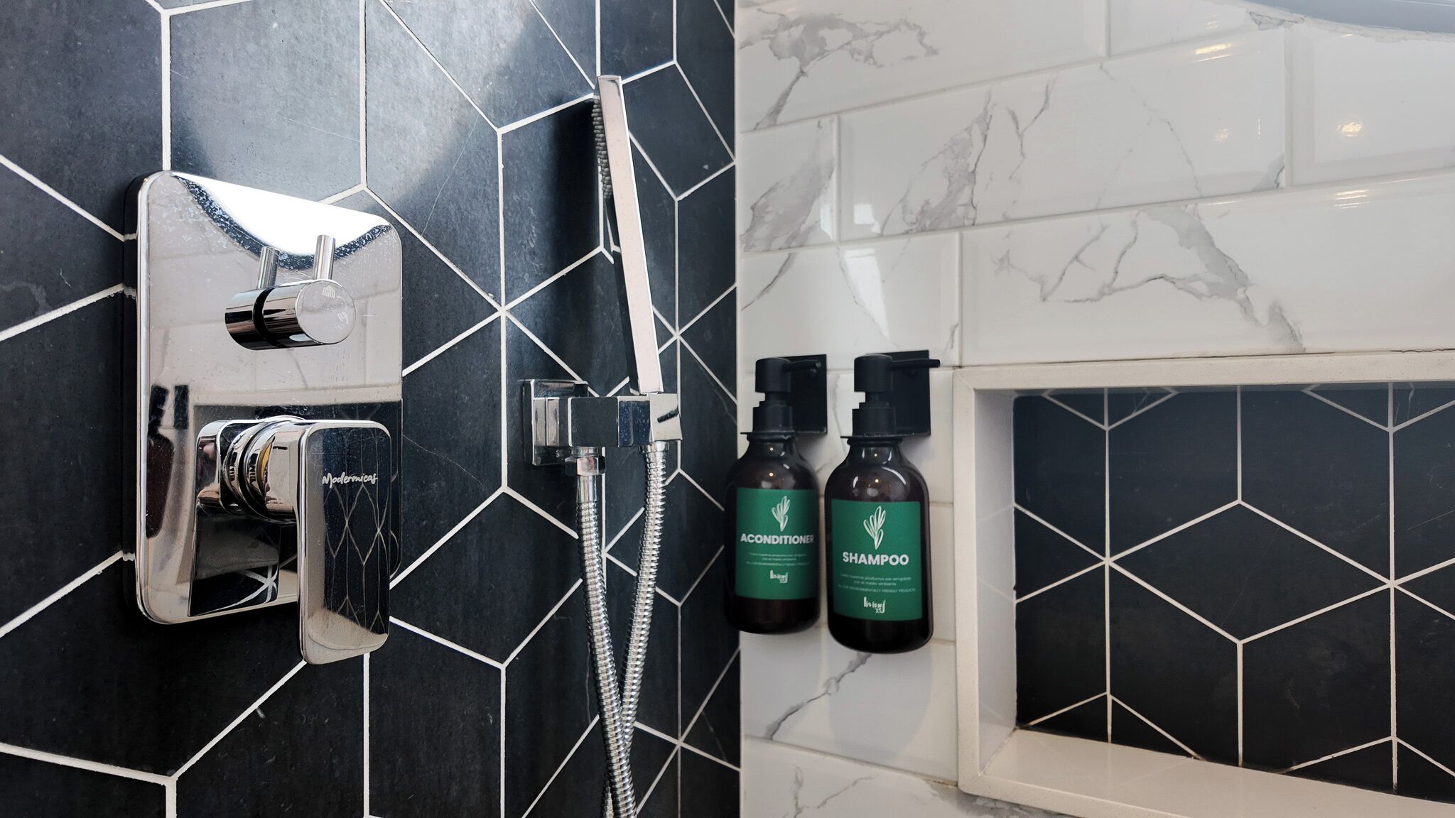 A bathroom with a modern design, featuring a shower with a handheld showerhead and a handheld faucet. There are two bottles of shampoo and conditioner on the wall.