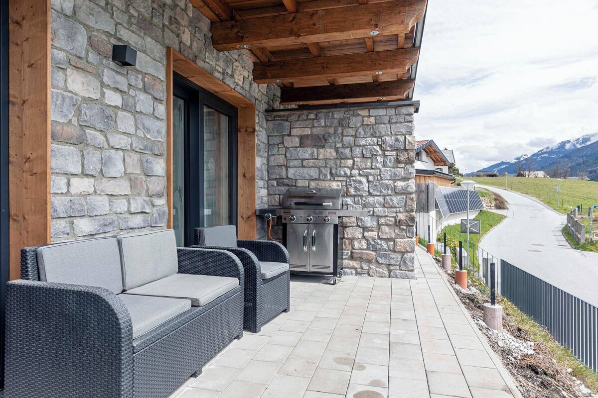 Fachada de piedra con un porche cubierto, mobiliario de exterior y una parrilla de barbacoa.