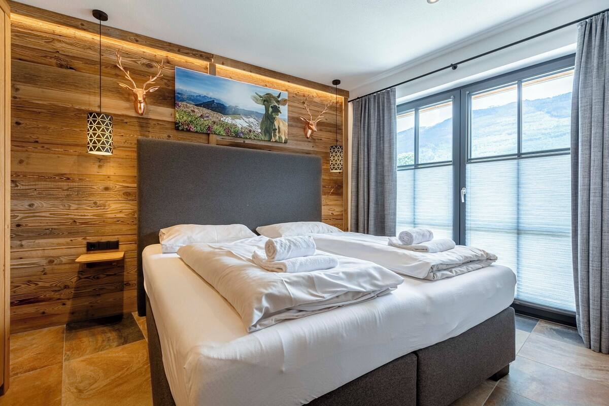 Un dormitorio moderno con una cama grande, paneles de madera en la pared, una cabeza de ciervo montada y una vista panorámica de la montaña a través de la ventana.