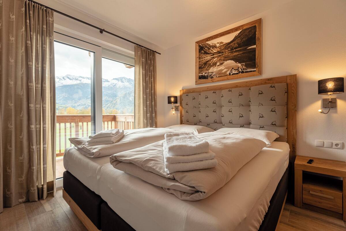 Habitación de hotel con una cama grande, un cabecero de madera, un cuadro sobre la cama, una mesita de noche y vistas a las montañas a través de la ventana.