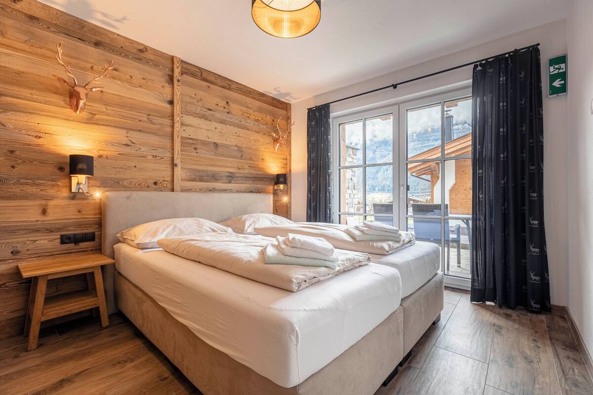 Un dormitorio con cabecera de madera, cama con ropa de cama blanca, mesita de noche y ventana con cortinas.