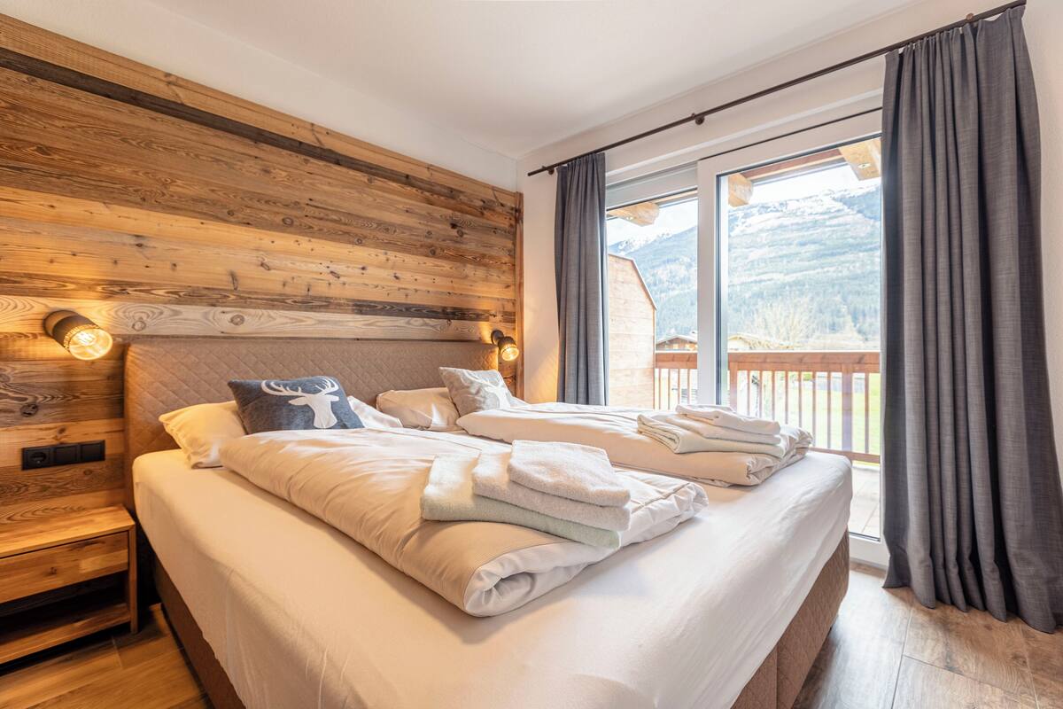 Habitación de hotel con pared de madera, una cama grande con ropa blanca, una mesita de noche y vistas a montañas a través de una ventana.