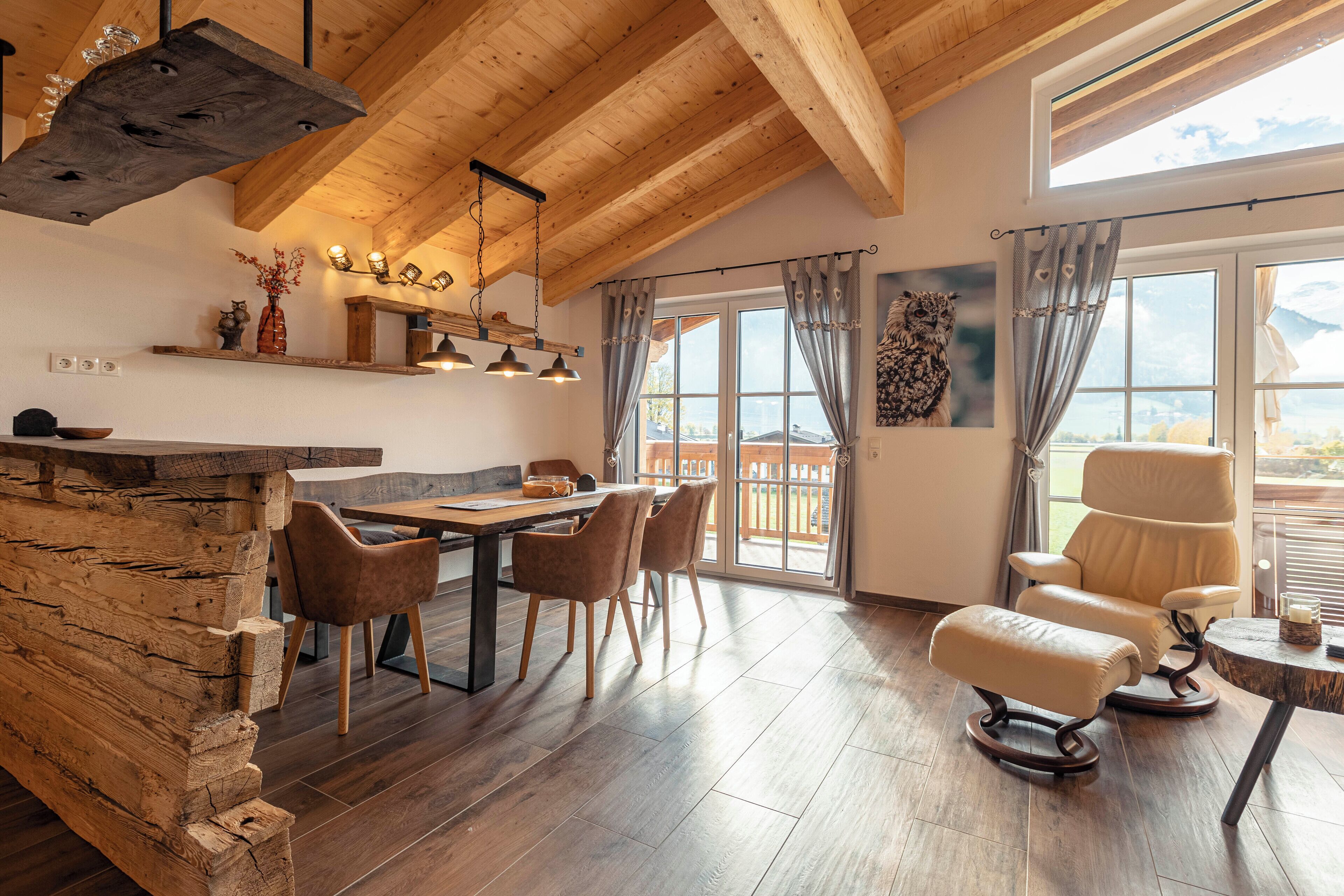 A spacious room with a wooden ceiling, a dining area with a table and chairs, a sofa, and a view of the outdoors through large windows.