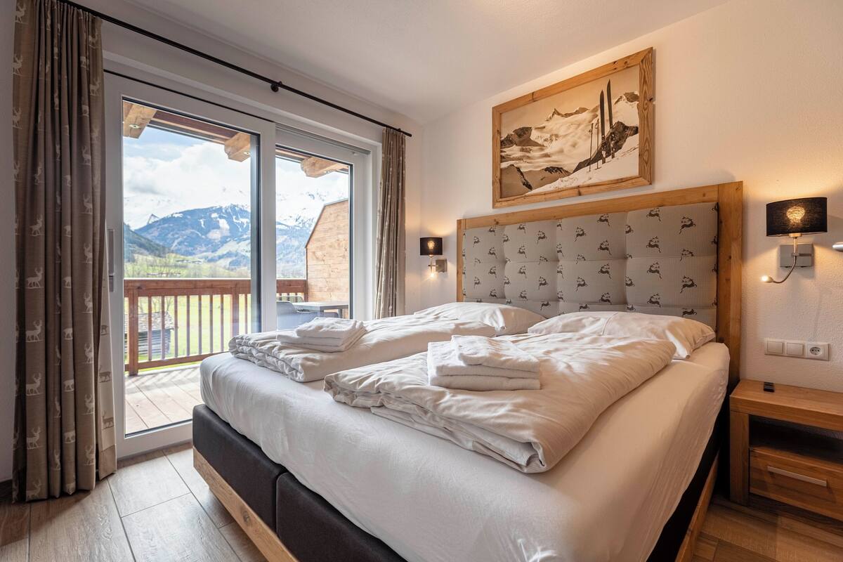 Un dormitorio con una cama grande, cabecera de madera, un cuadro de montañas, puerta corrediza de vidrio a un balcón y una mesita de noche con una lámpara.