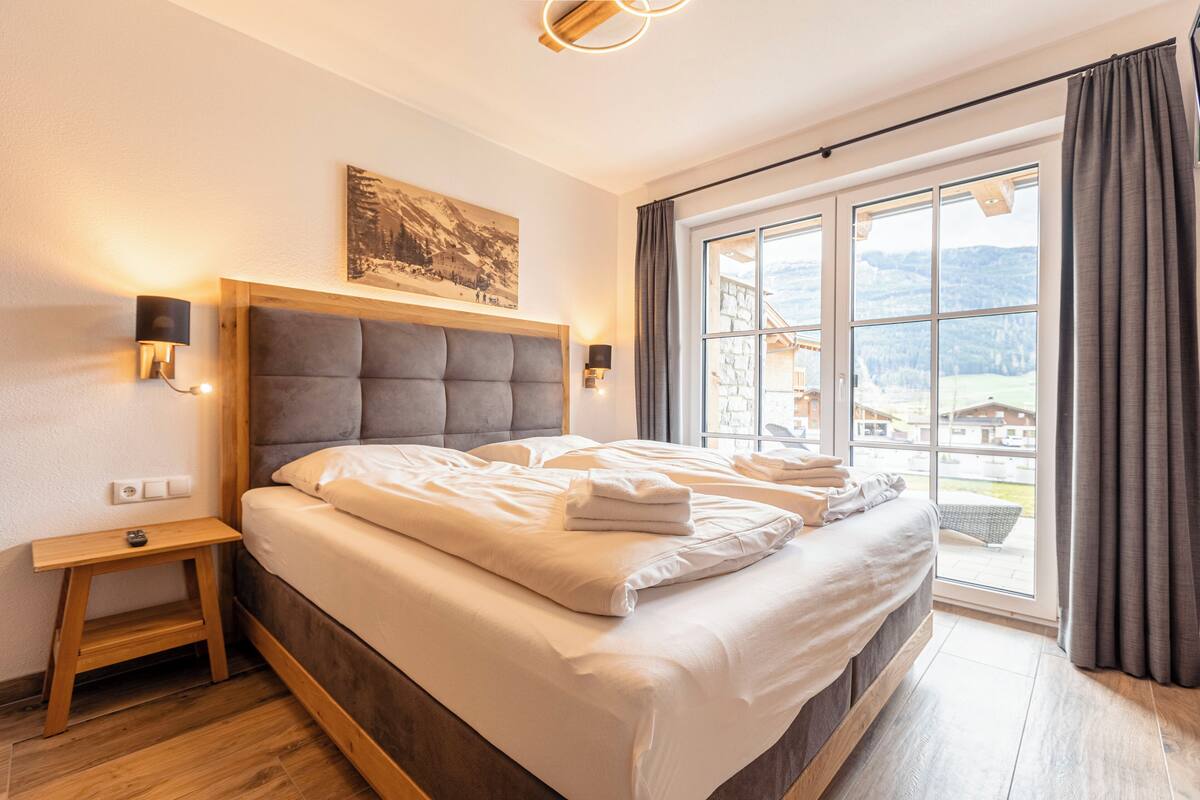 Un dormitorio con una cama grande, una mesita de noche, una lámpara de pared y una ventana con vistas a un paisaje montañoso.