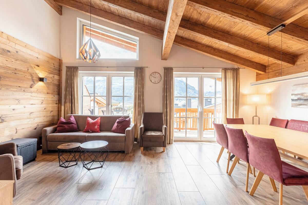 Un salón amplio con techo de madera, un sofá con cojines rojos, una mesa de comedor con sillas y grandes ventanales con vistas a la montaña.