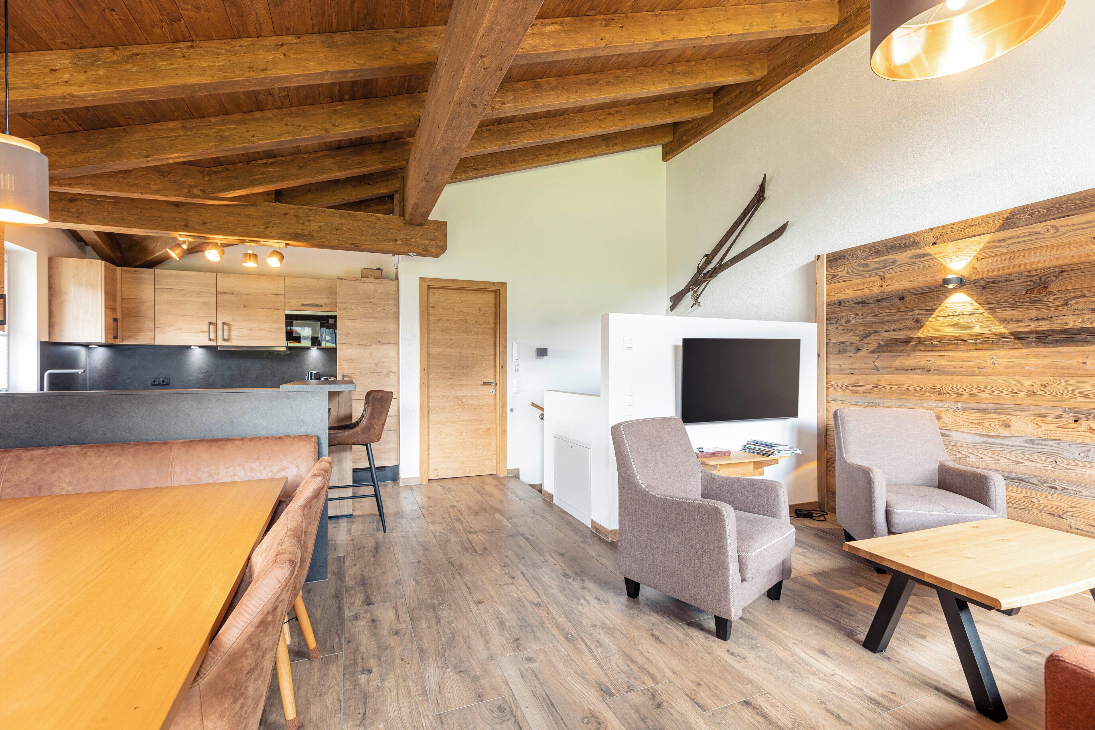 Una cocina moderna con vigas de madera, una zona de comedor con banco y un espacio de estar con televisor y asientos.