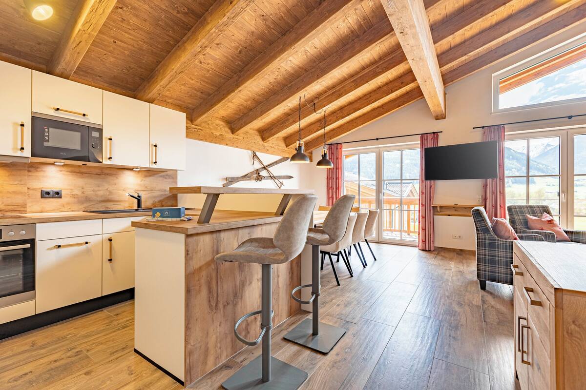 Una cocina moderna con vigas de madera, una zona de bar con taburetes, un televisor y vistas a montañas a través de grandes ventanales.