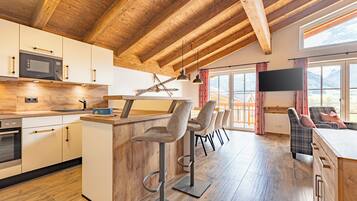 A modern kitchen with wooden beams, a bar area with stools, a TV, and a view of mountains through large windows.