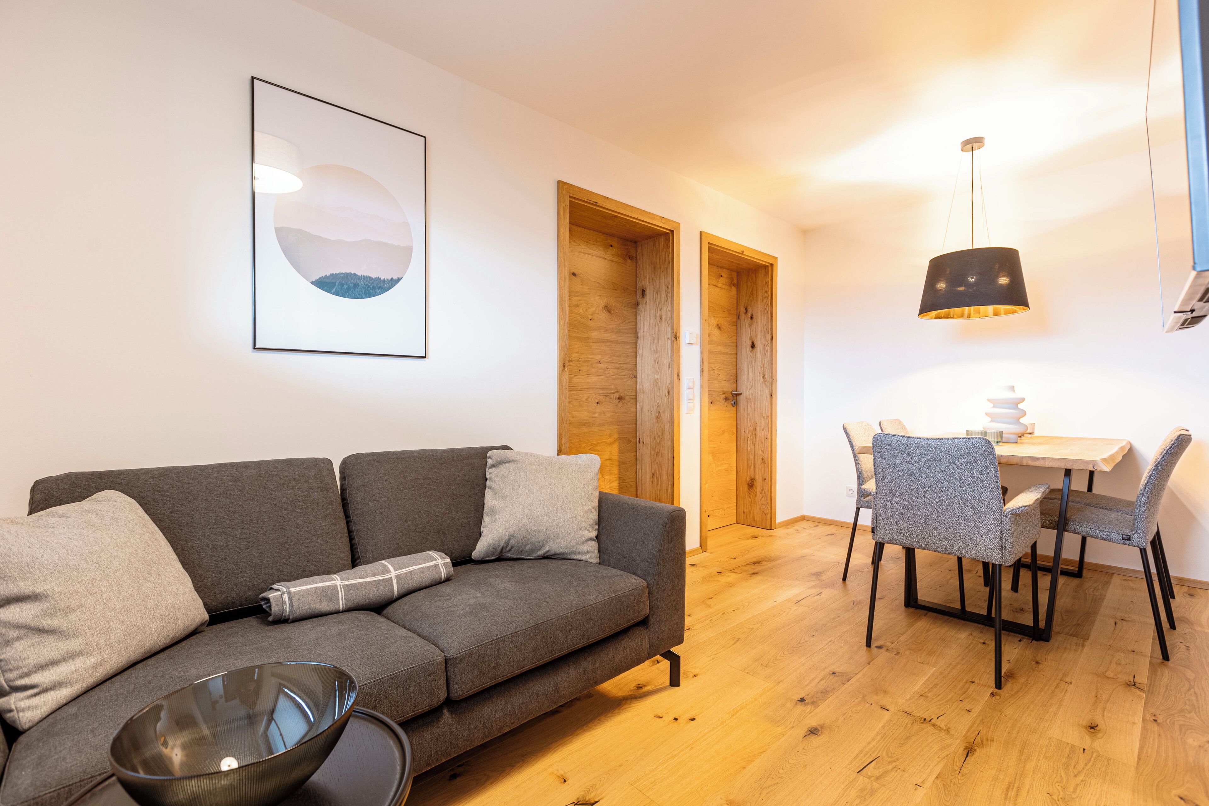 Un salon moderne avec un canapé gris, une table à manger avec des chaises, une porte en bois et un luminaire suspendu.