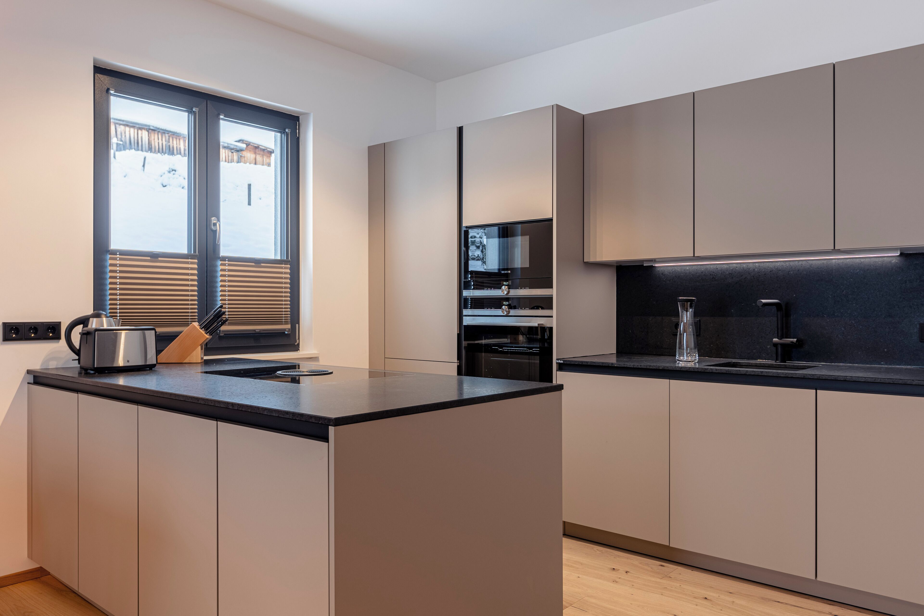 A modern kitchen with a central island, built-in appliances, and a window with blinds.