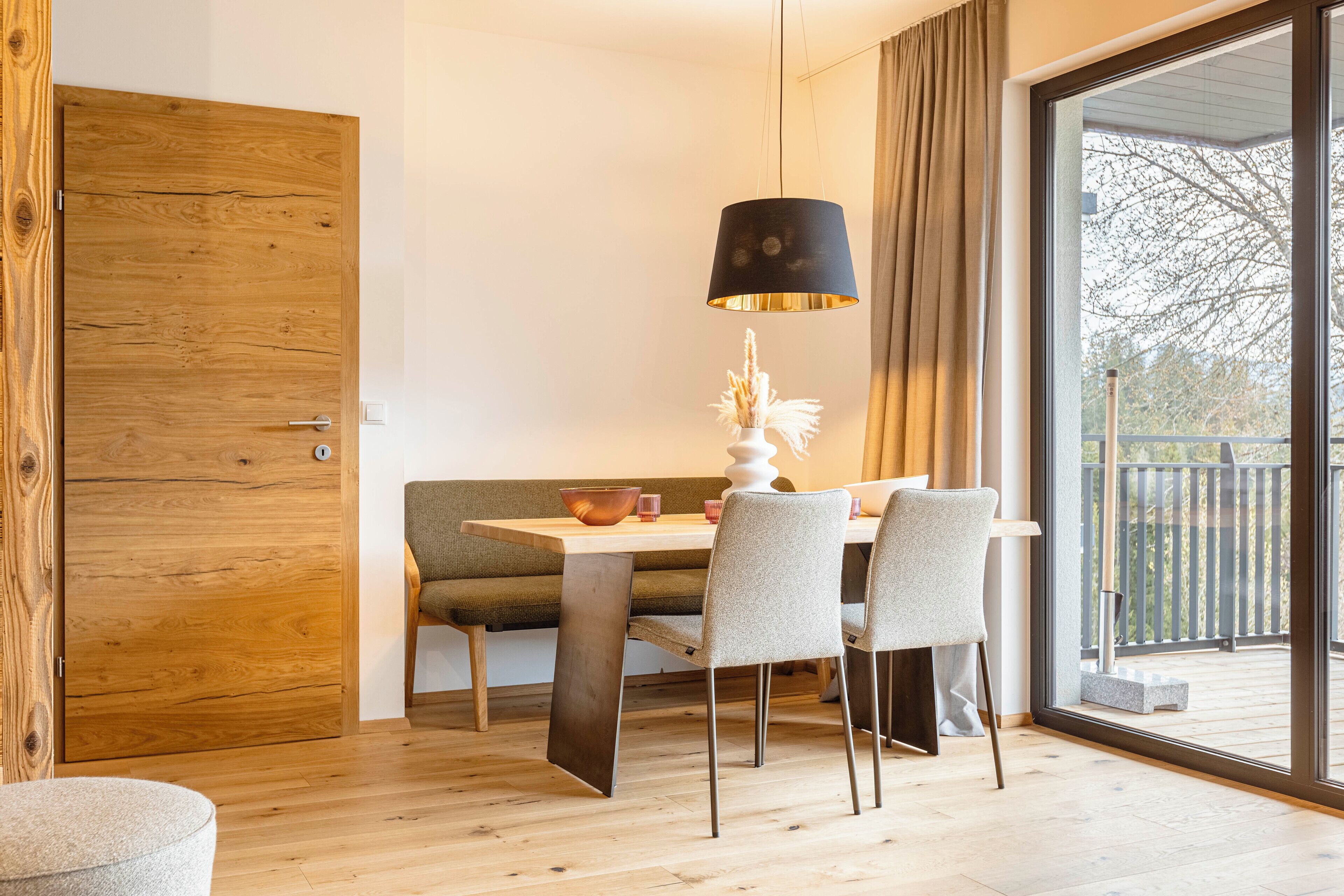 A dining area with a wooden table, chairs, a bench, and a large window with a view of a balcony.