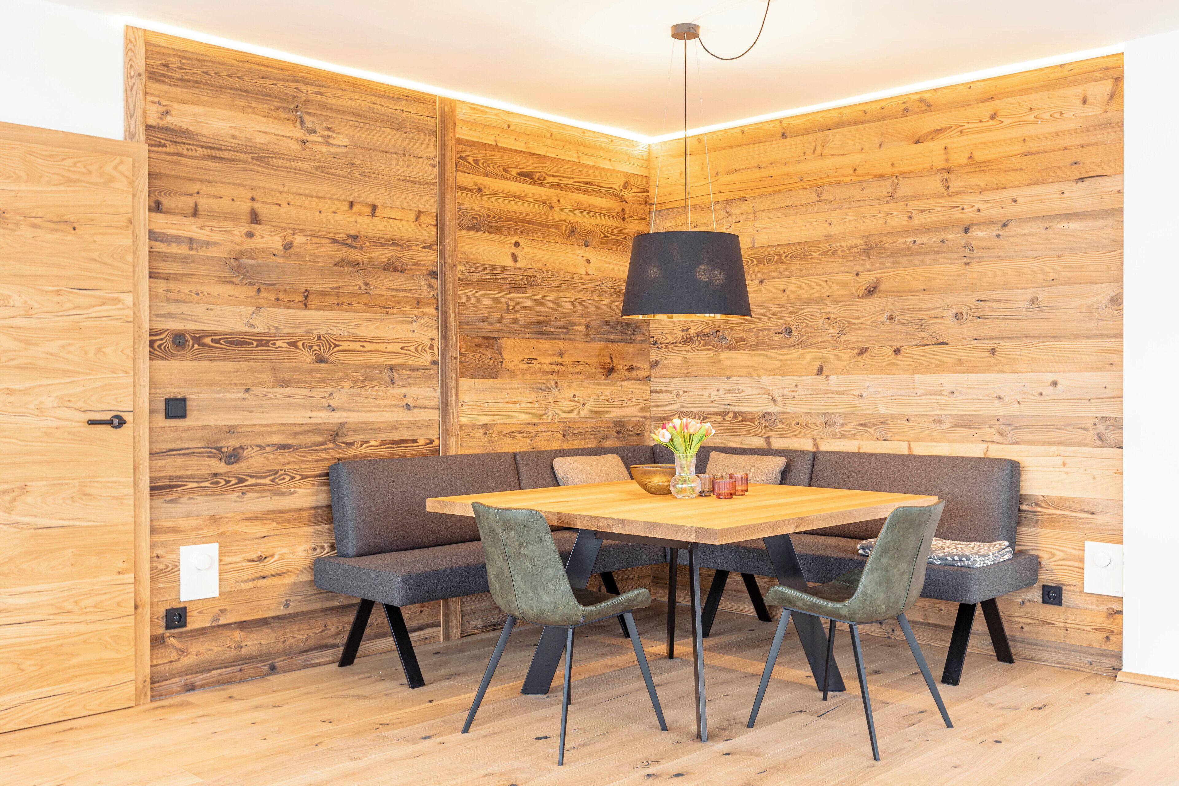 A room with a wooden wall panel design, a wooden dining table with four chairs, a sofa, and a hanging light fixture.