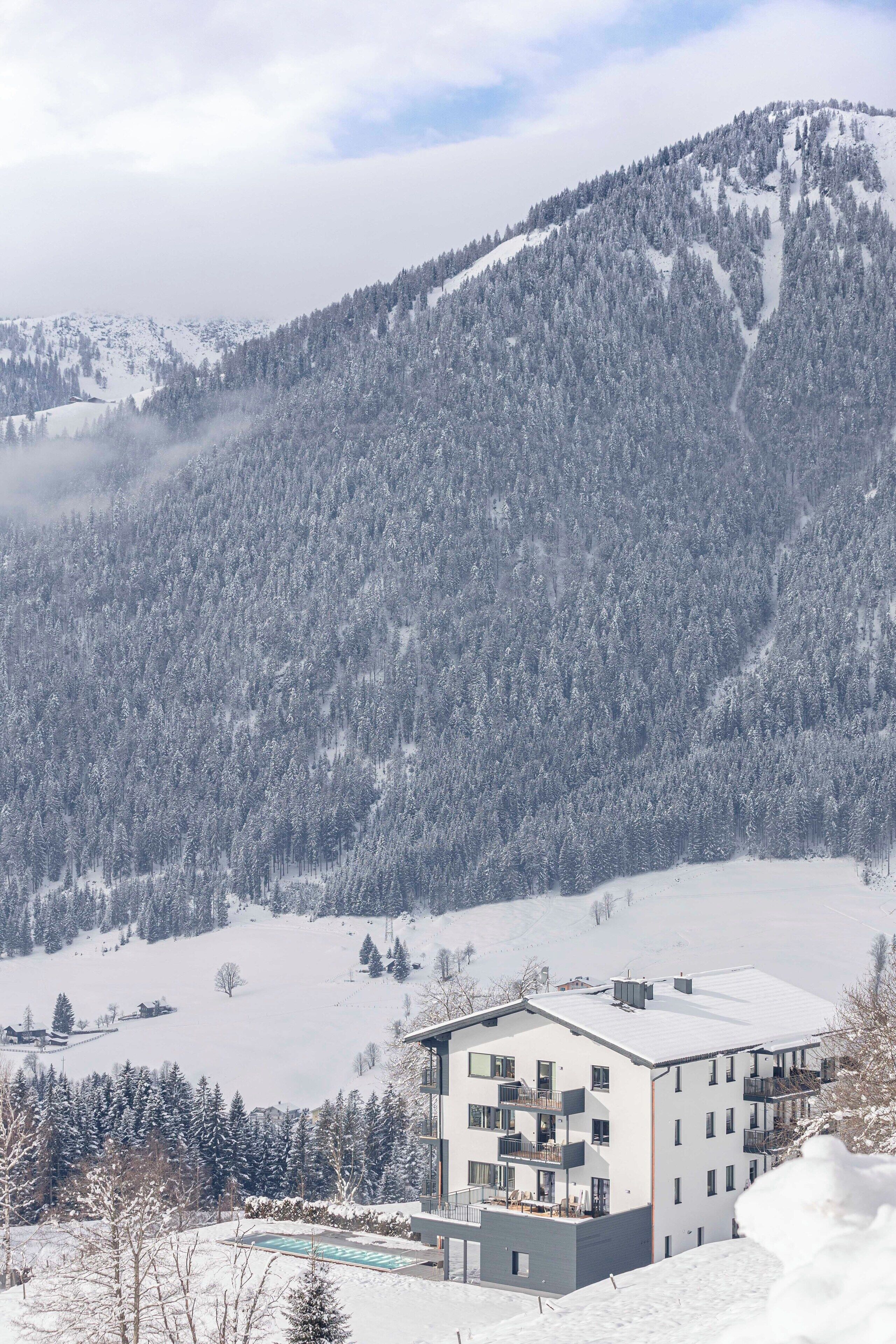 A modern multi-story hotel with a flat roof, surrounded by snow-covered mountains and trees.