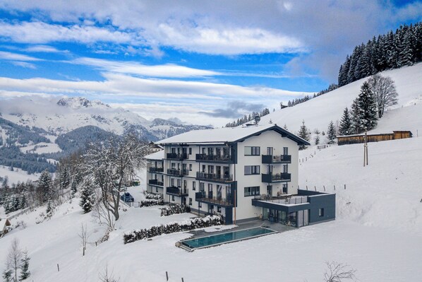 Un edificio moderno de varios pisos con balcones, piscina y una montaña nevada al fondo.