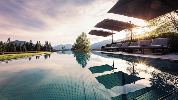Piscina límpida com espreguiçadeiras e guarda-sóis, cercada por árvores e montanhas.