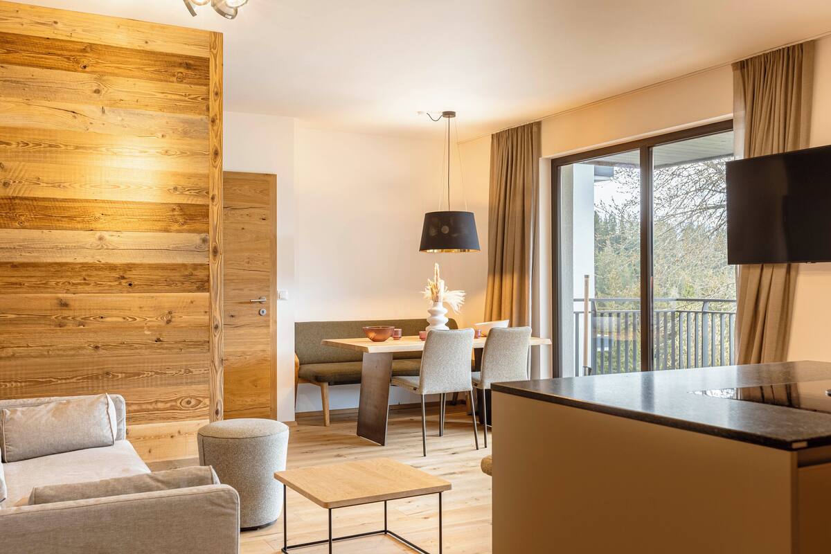Ein modernes Hotelzimmer mit einer Wandpaneele aus Holz, einer Essecke mit Tisch und Stühlen, einem Sofa, einem kleinen Hocker und einem an der Wand befestigten Fernseher.