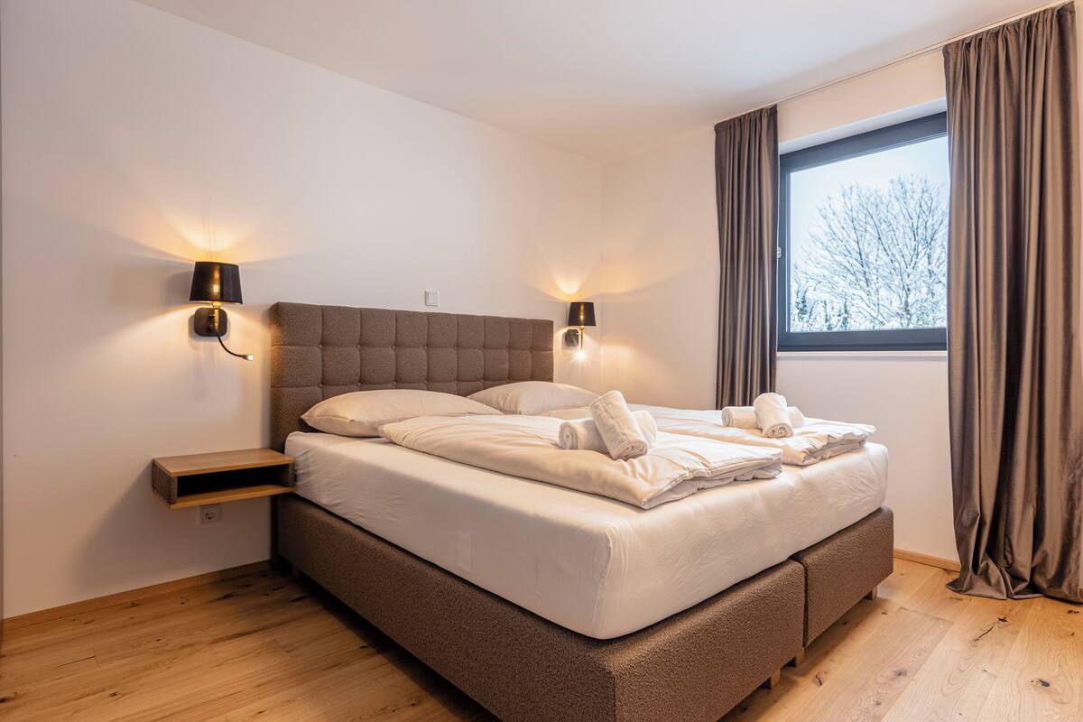 Ein modernes Schlafzimmer mit einem großen Bett, zwei Nachttischlampen, einem Nachttisch und einem Fenster mit Vorhängen.