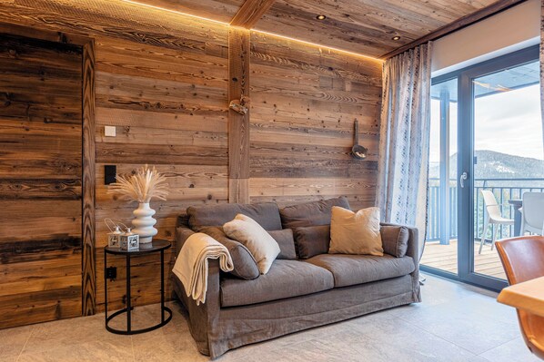 A cozy living room with a wooden wall, a grey sofa, and a small side table with a vase and a blanket.