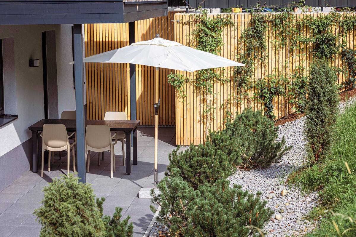 Eine Terrasse im Freien mit Tisch und Stühlen, einem weißen Sonnenschirm und einem Holzzaun mit Kletterpflanzen.