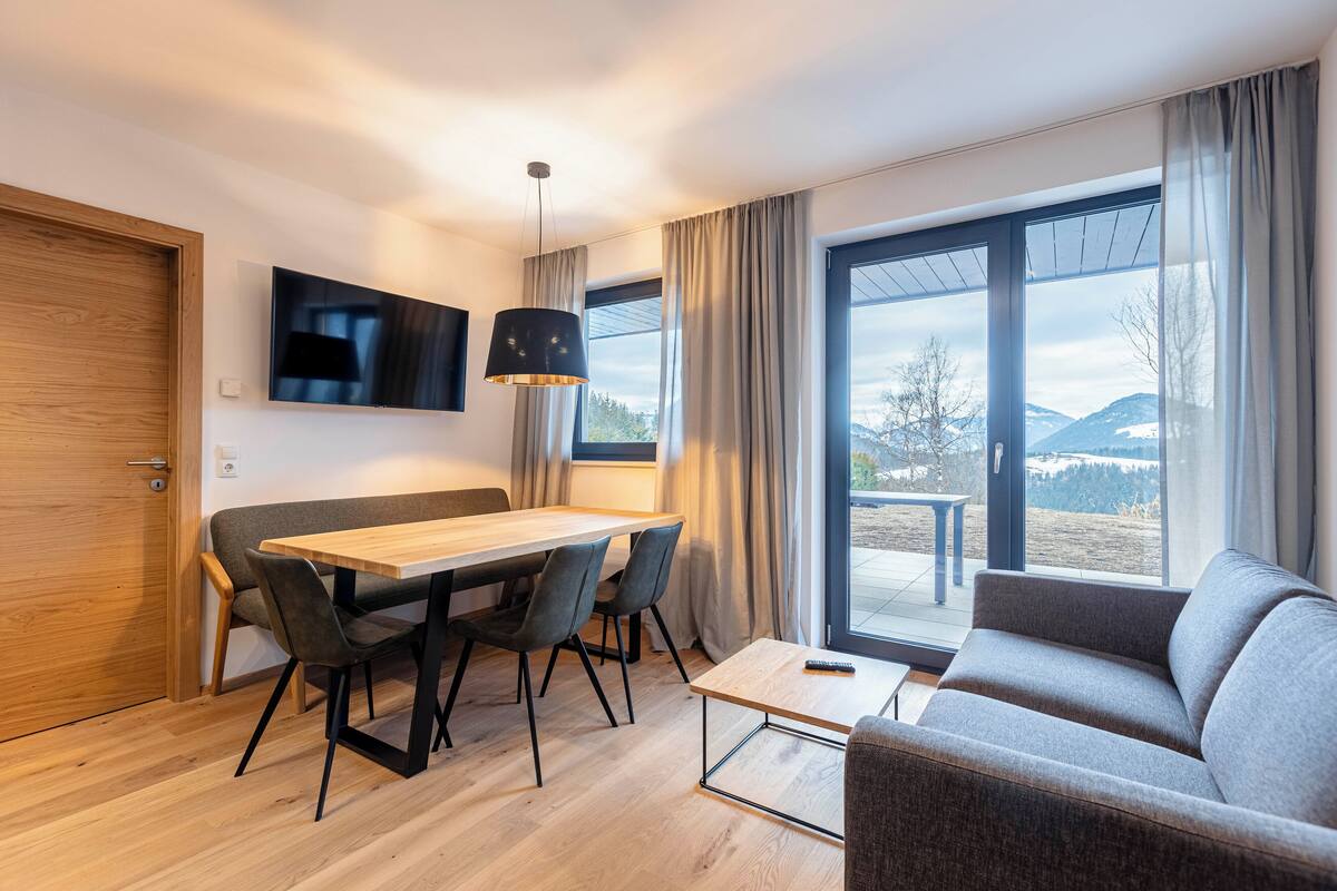 Ein modernes Wohnzimmer mit einem Holzesstisch, einer grauen Couch und einem an der Wand montierten Fernseher.